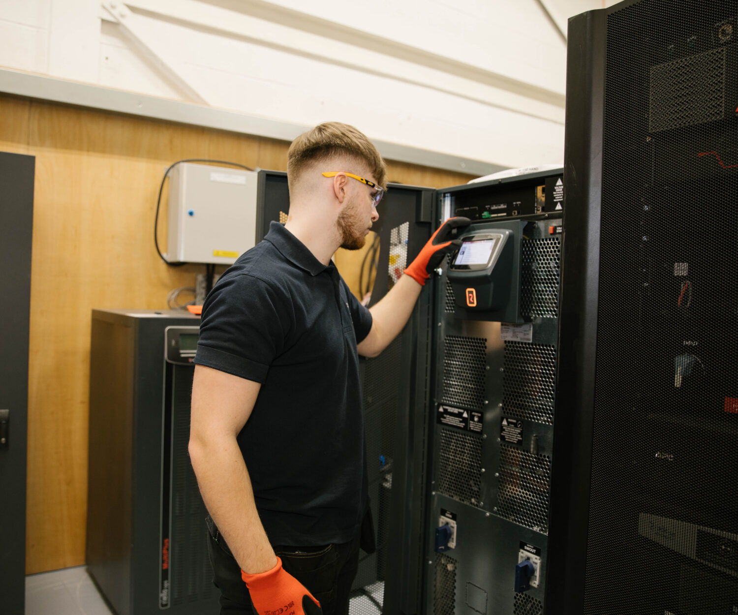 Man stood upright looking at power source wearing orange gloves